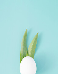 White egg and bunny ears made of green leaves on bright cyan background. Easter minimal concept. Flat lay.