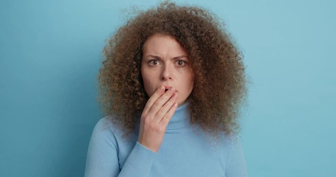 Stunned young woman stands speechless reacts amazed on unbelievable news covers mouth stands startled indoor says omg has curly bushy hair isolated over blue background. Human reaction concept
