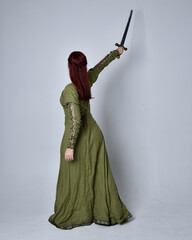 full length portrait of red haired girl wearing celtic, green medieval gown. Standing pose, holding a sword, isolated against a studio background.