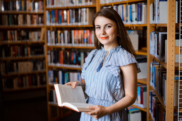 Smiling attractive woman with dark hair, in blue dress holding a book and looking to the camera. Girl in the library, looking for an interesting reading.
