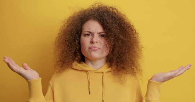 Lovely curly haired young woman raises palms shrugs shoulders feels doubt dressed in hoodie cannot make choice isolated over yellow background. Clueless European female face difficult choice