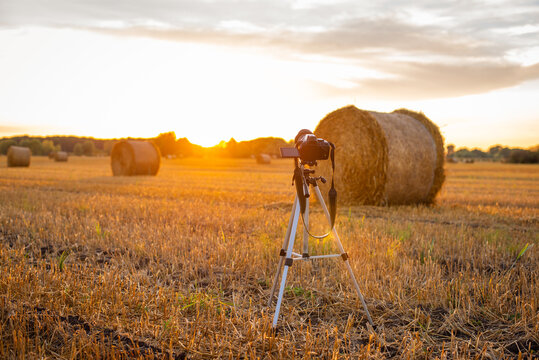 Evening in the farming field, sun over the horizon, straw bales after harvest. Professional camera on a tripod ready for taking photos.