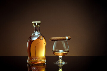 close up view of cigar, bottle of cognac and a glass aside on brown back. 