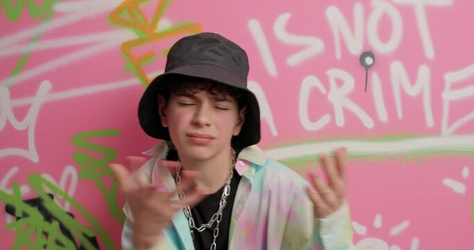 Handsome cool guy wears hat and metal chain makes rap gesture dances to favorite music against colorful painted graffiti wall. Urban lifestyle and activity concept. Fashionable youngster indoor