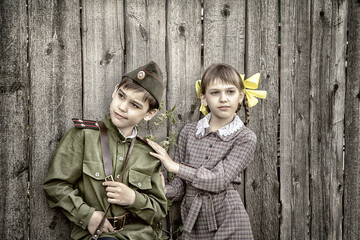 Postcard, stylized as vintage for the Victory Day. A boy in a military uniform and a girl in an old dress are standing near an old fence. The theme of May 9, Victory Day in Russia.