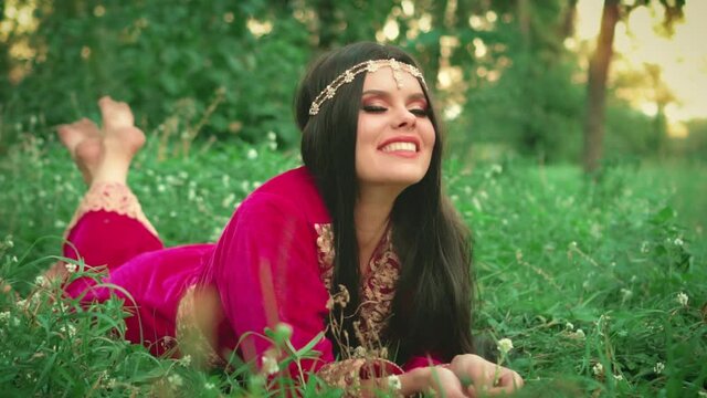 young happy oriental woman lies on green grass summer meadow. Hippie girl smiling beautiful face. Ethnic red dress golden headband on long black hair. cheerful joyful lady enjoying life freedom nature