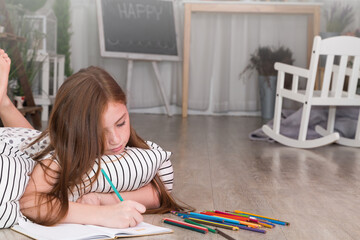 Portrait happy lovely daughter lying on floor enjoying drawing pictures in copybook and looking at camera. Happy with homework at home.