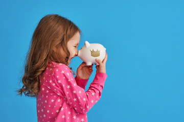 Happy savings. little cute girl with white piggy bank in her hands on blue background in studio....