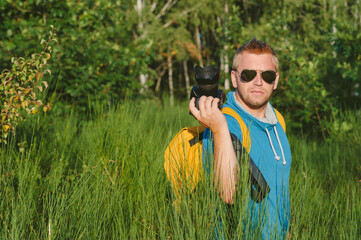 A man in a blue T-shirt and a yellow backpack holds a professional photo-video camera in his hands. Against the backdrop of green nature and forest.