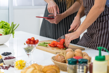 young caucasian woman chopping vegetable while husband using tablet to read instruction of how to cook healthy salad for breakfast in kitchen. couple together concept