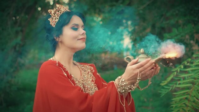 fantasy woman princess in red luxurious oriental dress holds in hand golden antique old lamp. Girl queen smiles. Mystical spirit, smoke magic miracle from a jug. Background summer forest green tree 