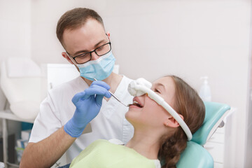 Professional dentist examining teeth of young girl, using inhalation sedation mask on her