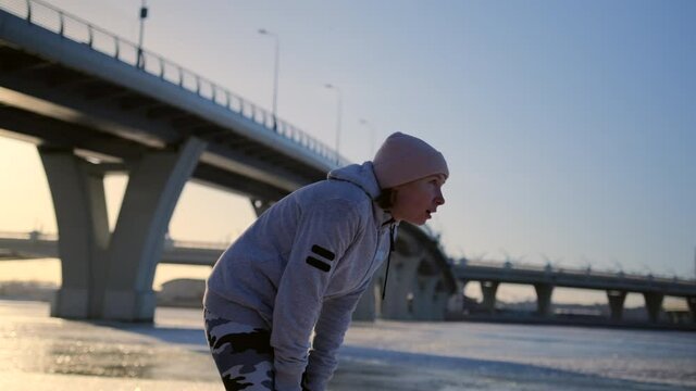 The runner finished an arduous workout under a bridge next to a frozen river in winter. The girl finished a grueling run took off her hat and took a breath. Tired girl after jogging