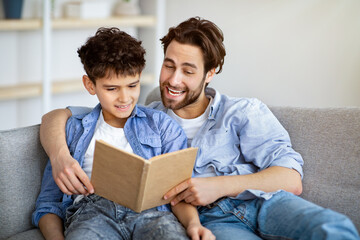 Useful time at home. Father and son reading book and smiling, resting together on comfy sofa.