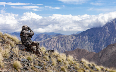 Man in camouflage on the slope is watching through binoculars in the mountains.