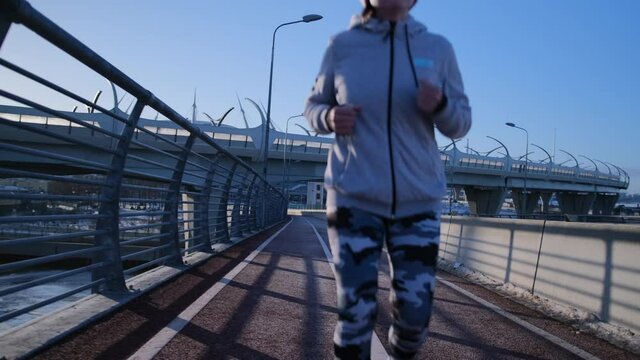 The girl runs back across the bridge over the river in winter. A girl runs backwards on a treadmill early in the morning in camouflage leggings and warm clothes