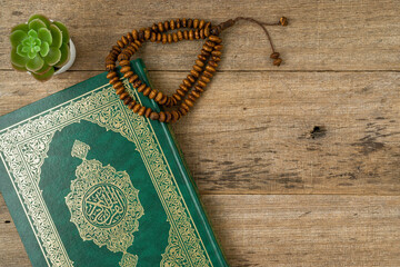 Holy Al Quran with written arabic calligraphy meaning of Al Quran and rosary beads or tasbih on wooden board background with a copy space. Flat lay.