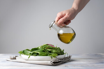 Female hand pouring olive oil into a plate with a salad of leafy greens on white wooden background, healthy eating
