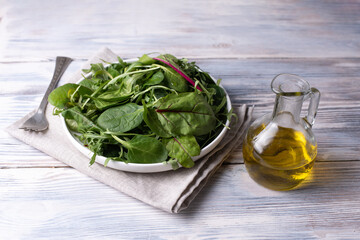 Plate with fresh leafy greens, bottle of olive oil and fork on white wooden table, vegetarian food,...