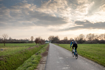 Outdoor sunny view of cyclist ride a bicycle on small road in suburb area surrounded with agricultural field in spring season against dramatic sky in Germany.