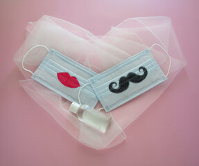 Two disposable protective masks with mustache and lips painted on them and two wedding rings on heart-shaped veil on pink background. The concept of wedding ceremonies during the coronovirus epidemic