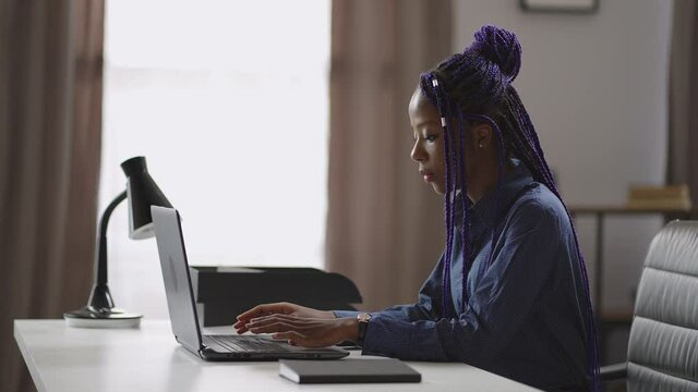 adult black woman is learning online in home, sitting at table with laptop, typing on keyboard, portrait