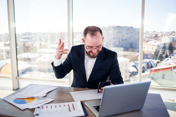 Fototapeta premium Adult successful male businessman, teacher, mentor is working on a new project. Sits at the table by the large window. It works on a laptop