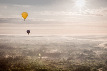 ein gelber und ein dunkler Heißluftballon fliegen über einer dunstigen Landschaft in der frühen...
