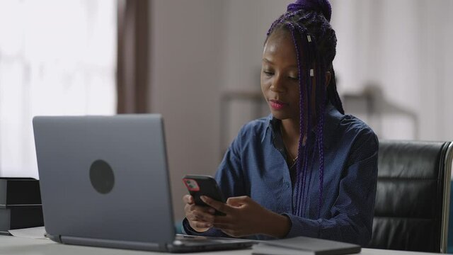 afro-american lady is using smartphone during working day in office, checking email and message in social nets, sitting at table with laptop