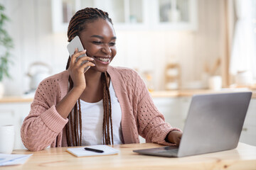 Remote Business. Smiling African Freelancer Lady Using Cellphone And Laptop In Kitchen