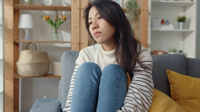 Thoughtful Asia lady sit embracing knees at sofa in living room at house look outside with feel lonely, Sad depressed teenager spend time alone stay at home, Social distance, Coronavirus Quarantine.