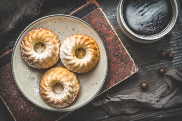 Three homemade mini bundt cakes on a plate, a cup of coffee and an old book on wooden background, top view