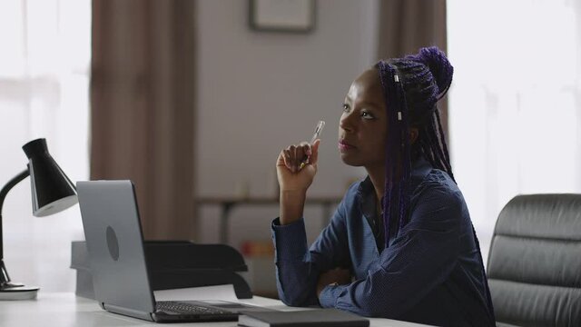 female writer is working over new book or article, afro-american woman sitting alone in room in apartment, holding pen and thinking