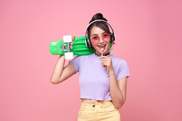 Young asian teenage girl holding skateboard with lollipop on pink background.