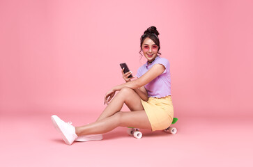 Young asian teenage girl sitting on skateboard and holding smartphone on pink background.