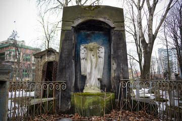 Stone crypt with headless statue of angel and metal fence on background of urban houses 