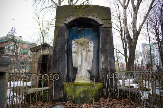 Stone crypt with headless statue of angel and metal fence on background of urban houses 