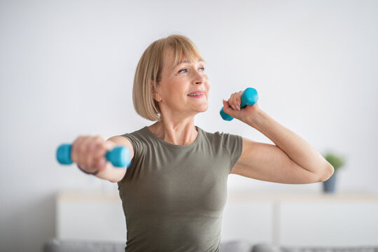 Domestic sports training. Positive senior woman doing exercises with dumbbells, strengthening her body at home