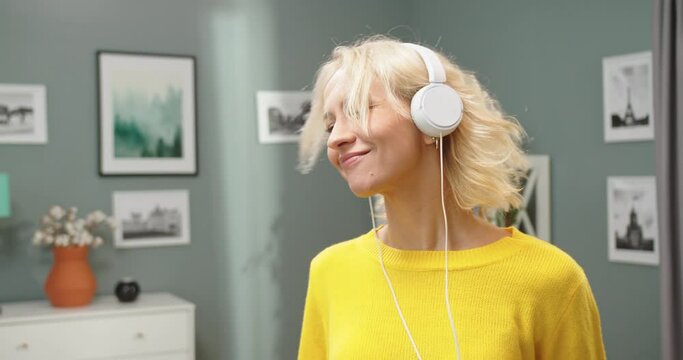 Close up of joyful relaxed young Caucasian beautiful blonde woman dancing and moving in cozy living room in house while listening to music in headphones having fun enjoying fav song
