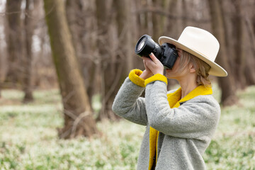 stylish girl in gray coat with a camera in snowdrops meadow in forest