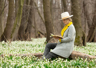 stylish girl in gray coat with a book in snowdrops meadow in forest