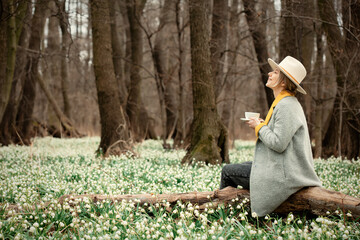 stylish girl in gray coat with a cup in snowdrops meadow in forest