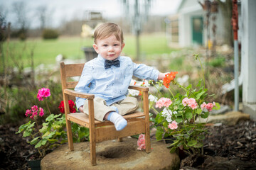 Cute baby boy in bowtie sitting outdoors with flowers on farm celebrating spring or Easter or Mother's Day