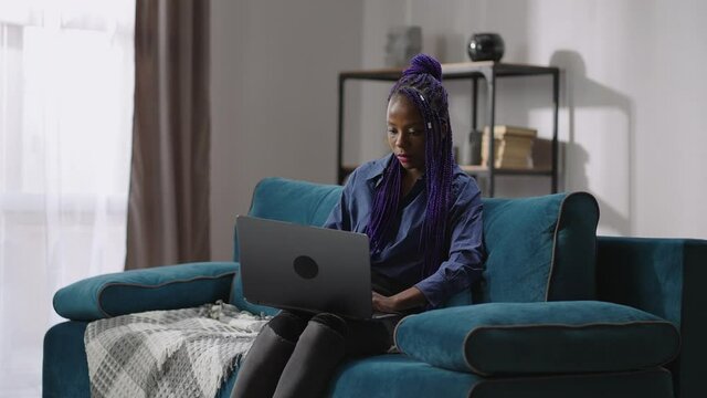 young black woman with purple dreadlocks is working remotely with laptop at home at weekend, freelance job for student