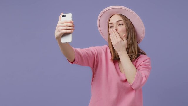 Cheerful blonde young woman in pink t-shirt hat isolated on violet purple background studio. People lifestyle concept. Doing selfie shot on mobile phone blowing send air kiss put hand on head cheek