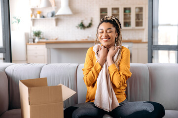 Happy satisfied stylish african american girl with dreadlocks, blogger, sits at home, in casual trendy wear, unpacks the parcel, hugging a new clothe, closes her eyes, smiling, happy with new purchase