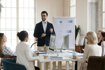 Smiling young Caucasian male coach or trainer make flip chart presentation for diverse colleagues...