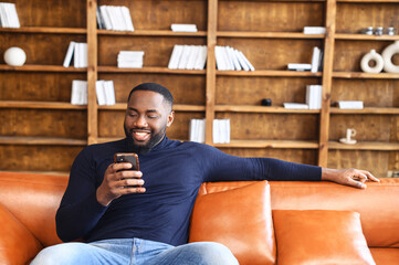 Portrait of smiling african american man sitting on sofa in office, using mobile phone for checking social nets, typing message for family, girlfriend, business partner, views photos, watches videos