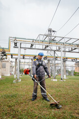 A young man mowing grass on the territory of an electric substation in overalls. Grass cleaning at the enterprise, implementation of fire safety measures