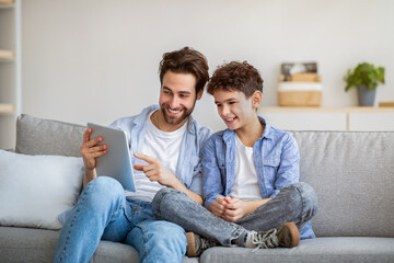 Joyful father and son using tablet computer, watching videos or playing online games, spending time...